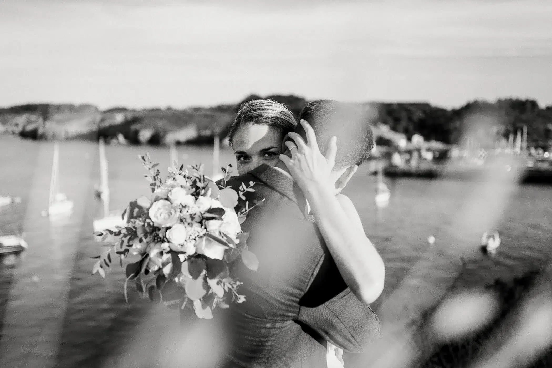 Couple de mariés s'enlaçant, mariée regard caméra, bouquet à la main, devant un port de voiliers à Belle Ile.