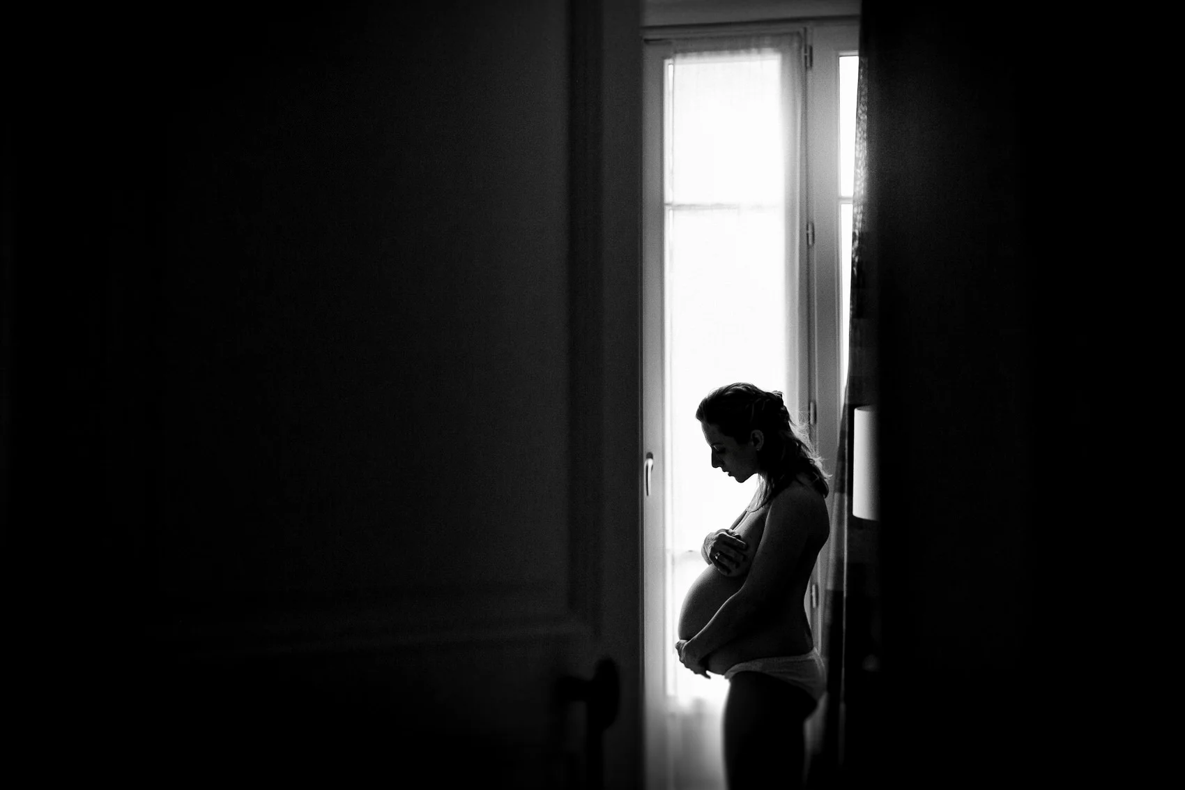 Séance photo grossesse à domicile, silhouette de femme enceinte en contre-jour près d'une fenêtre, noir et blanc