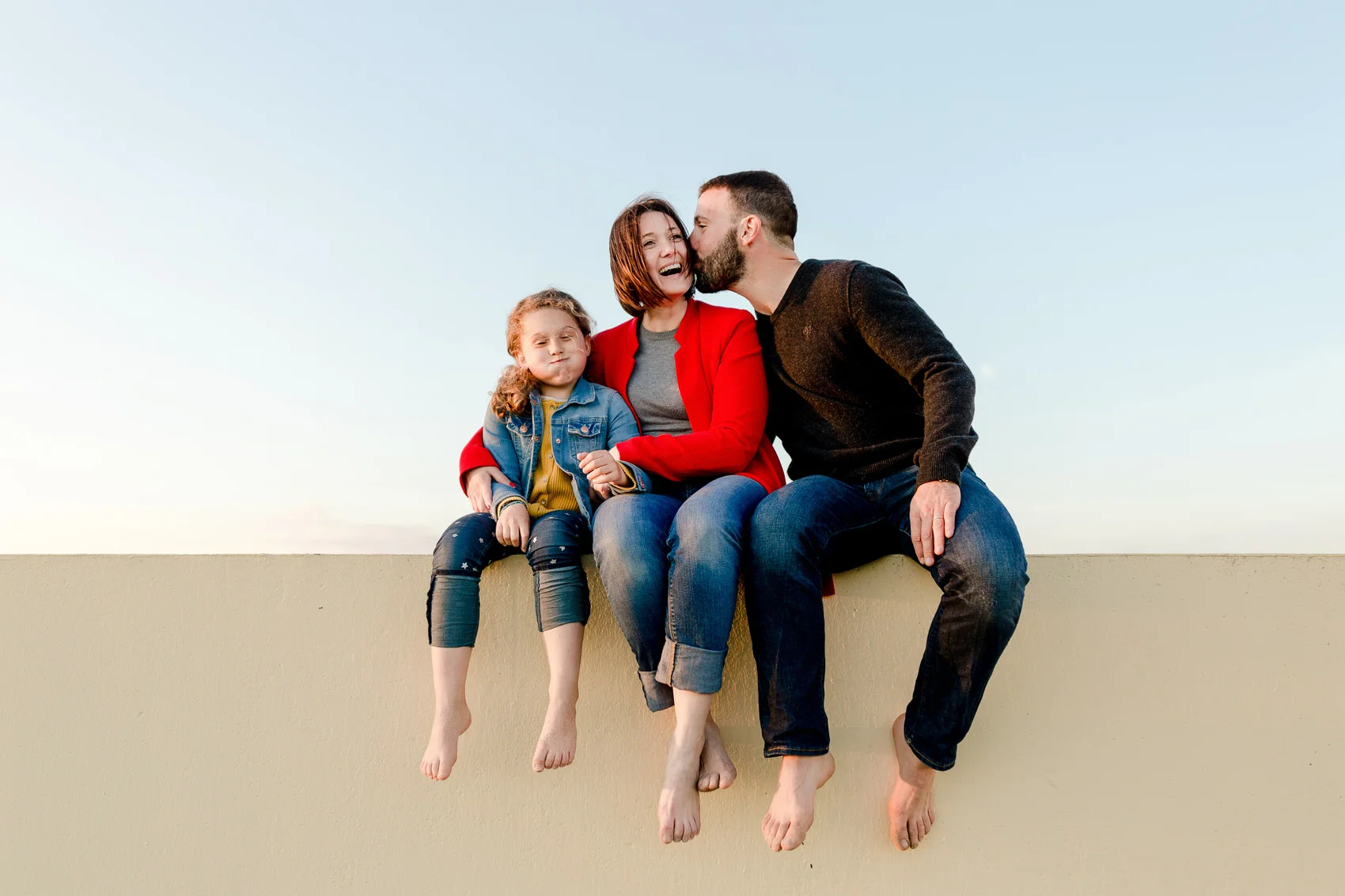 Séance photo famille à Arzon, parents et fillette assis pieds nus sur un muret, grimace et éclats de rire