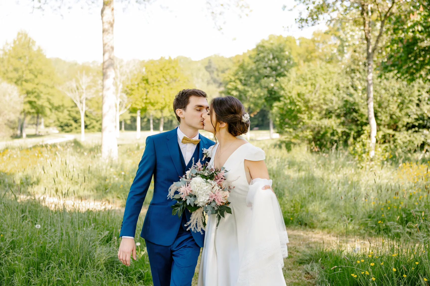 Couple de mariés s'embrassant en marchant dans un parc arboré à Vallet, marié en costume bleu, mariée avec bouquet champêtre